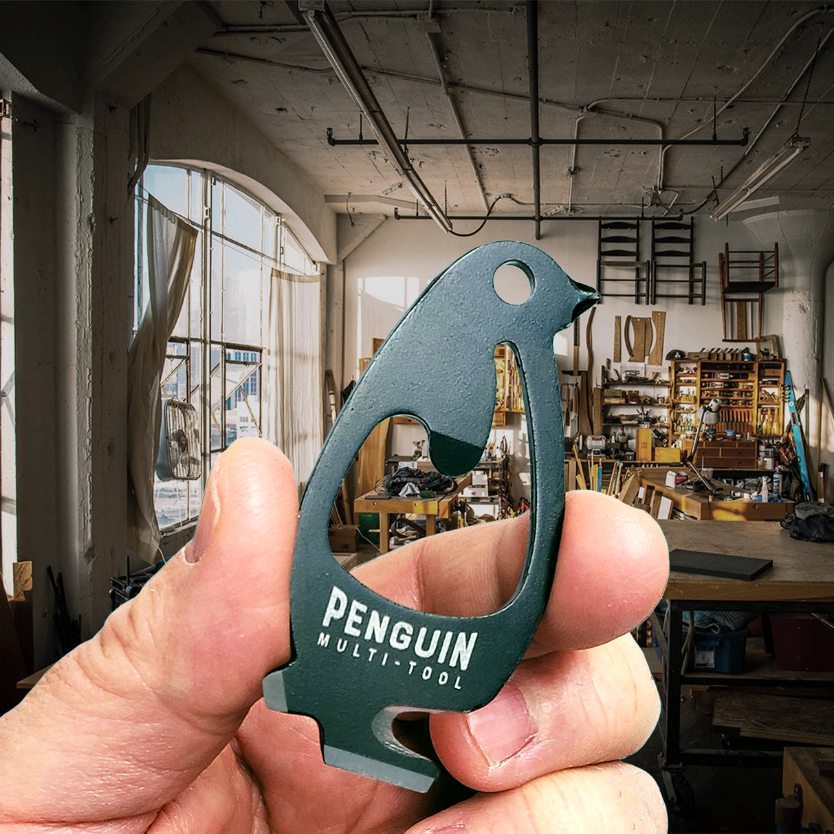 penguin tool in a workshop in front of a work bench. stainless steel coated in matte black pocket multitool shaped like penguin includes line cutter, scraping blade, bottle opener and more.